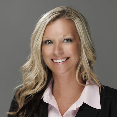 Nikki Pursell, Smiling woman with long blonde hair wearing a black blazer and pink blouse against a plain gray background, conveying a professional and friendly tone.