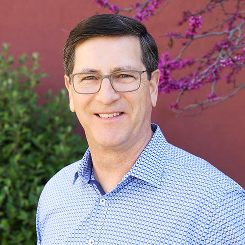 Tom Page, a smiling man wearing glasses and a blue patterned shirt stands in front of a red wall with green bushes and purple flowers, exuding a cheerful vibe.