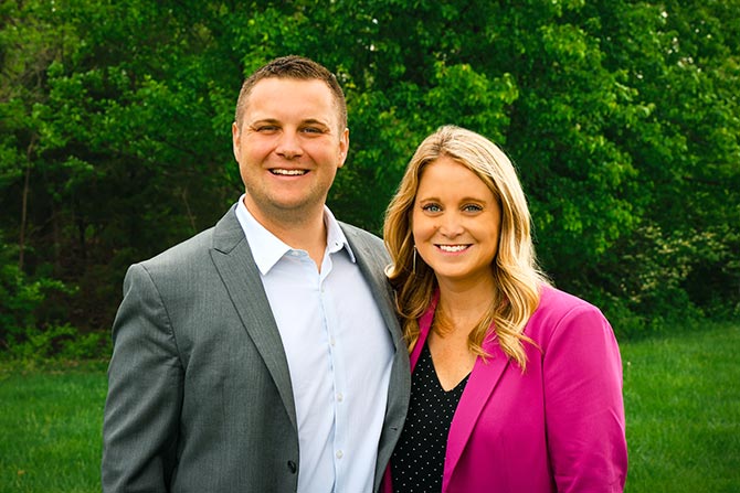 Kevin and Lynsey, a smiling man in a gray suit and woman in a pink blazer stand together outdoors, with green trees in the background, conveying a cheerful atmosphere.