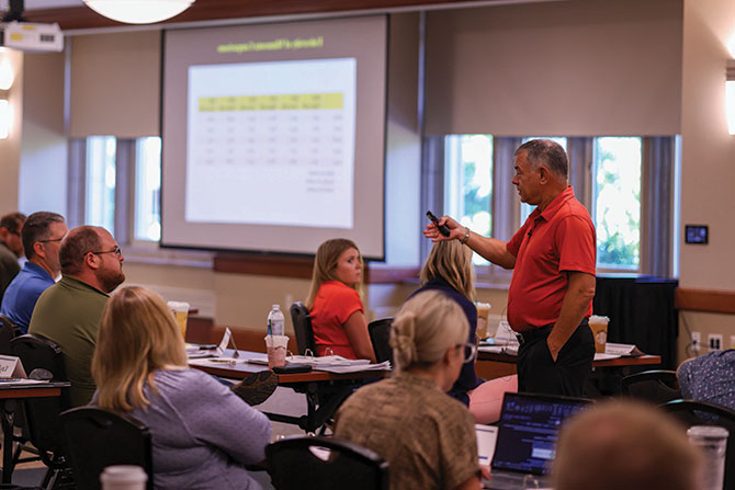 Agricultural Lenders School; A man in a red shirt speaks to seated attendees in a conference room, with a presentation slide showing data displayed on a screen.