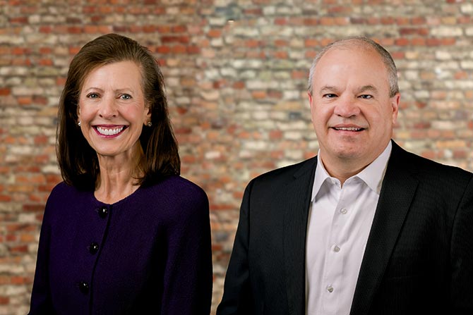 Background On BCC; Two people are smiling in front of a reddish-brown brick wall. The woman wears a dark purple jacket, and the man wears a dark suit with a white shirt.
