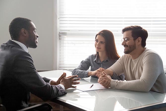 Financial Education; A confident businessman in a suit converses with a smiling couple across a table in a bright office. Papers lie on the table, indicating a discussion.