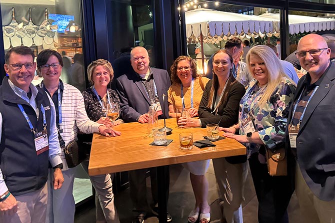 ICBA LIVE State Reception; Eight people are smiling around a tall wooden table at an indoor gathering, holding drinks. The atmosphere is relaxed and social, with warm lighting.