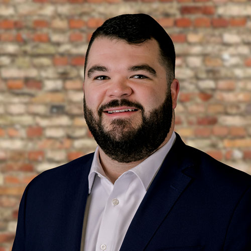 Kaleb Earls, a smiling man with a beard is wearing a dark suit and white shirt. The background is a rustic brick wall, providing a warm, professional tone.