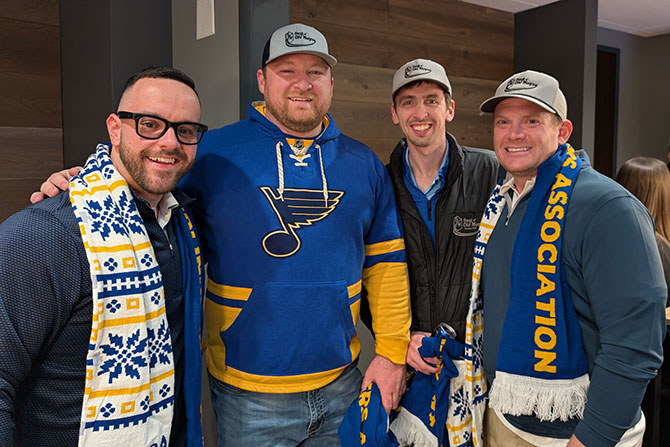 Leadership Division Day Out at the Blues; Four smiling men wearing hats and team scarves pose for a photo. One man in a blue and yellow sports jersey stands out. The mood is cheerful and camaraderie is evident.