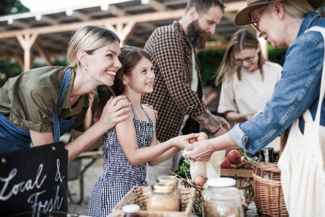 President’s Message Neighbors Helping Neighbors; A cheerful market scene with a mother and daughter buying fresh produce from a vendor. They're surrounded by baskets of fruits and jars, evoking a lively, local atmosphere.