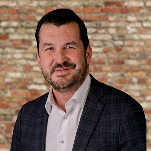 Rob Barton, a man in a dark plaid suit jacket and white shirt smiles subtly against a textured brick wall background. The tone is professional and approachable.
