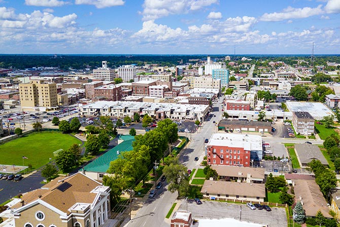 From the Top Your Work to Support the Future of Community Banking; Aerial view of a vibrant cityscape under a partly cloudy sky, showing diverse buildings, green spaces, and neatly lined streets, conveying urban vitality.